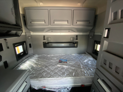 Truck sleeper cab interior with storage cabinets above and around a wrapped mattress in a Mack AN64T70 sleeper, compact dashboard area visible.