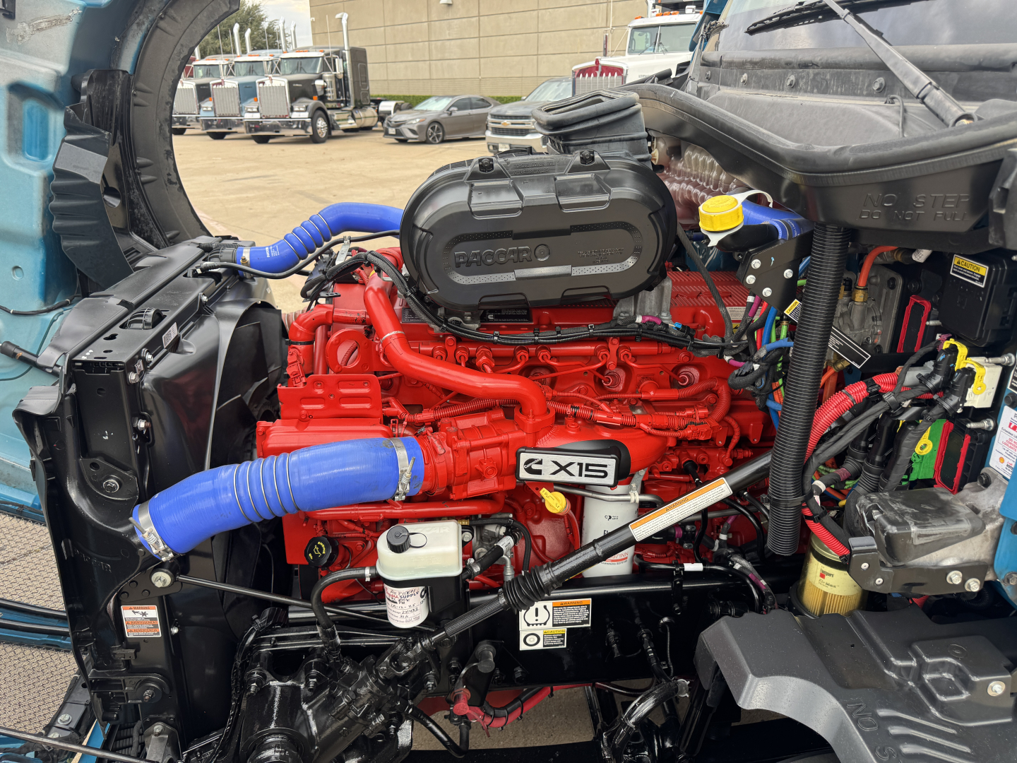 Kenworth T680 engine bay showing red Cummins ISX15 engine with blue hoses, black air filter housing, and various wiring; reservoir caps visible.