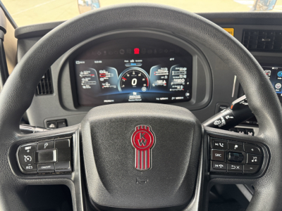 Kenworth T680 cockpit steering wheel with digital instrument cluster visible behind; dashboard controls on left and right, Kenworth emblem centered on the wheel.