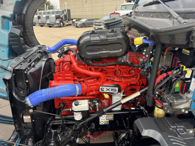 Kenworth T680 engine bay showing red Cummins ISX15 engine with blue hoses, black air filter housing, and various wiring; reservoir caps visible.