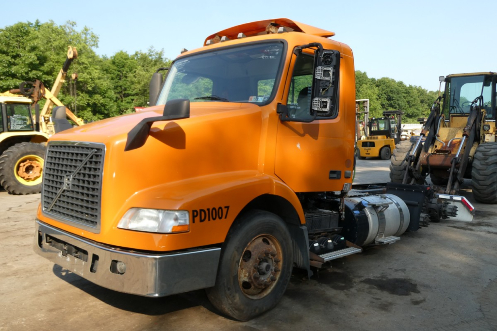 Orange Volvo VNM64T daycab truck side view showing PD1007 on door and fuel tank under cab with heavy equipment in background.