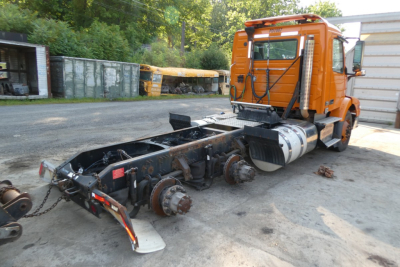 Truck chassis with cab and exposed drivetrain; orange Volvo cab in background; rear axle and suspension components visible, no wheels attached.