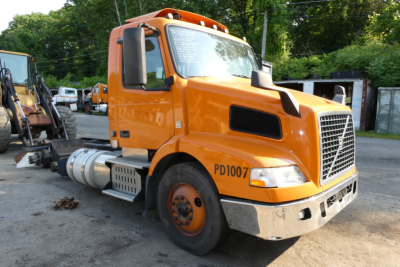 Orange Volvo VNM64T truck cab with PD1007 markings, front grille, diesel fuel tanks, and side steps in yard.
