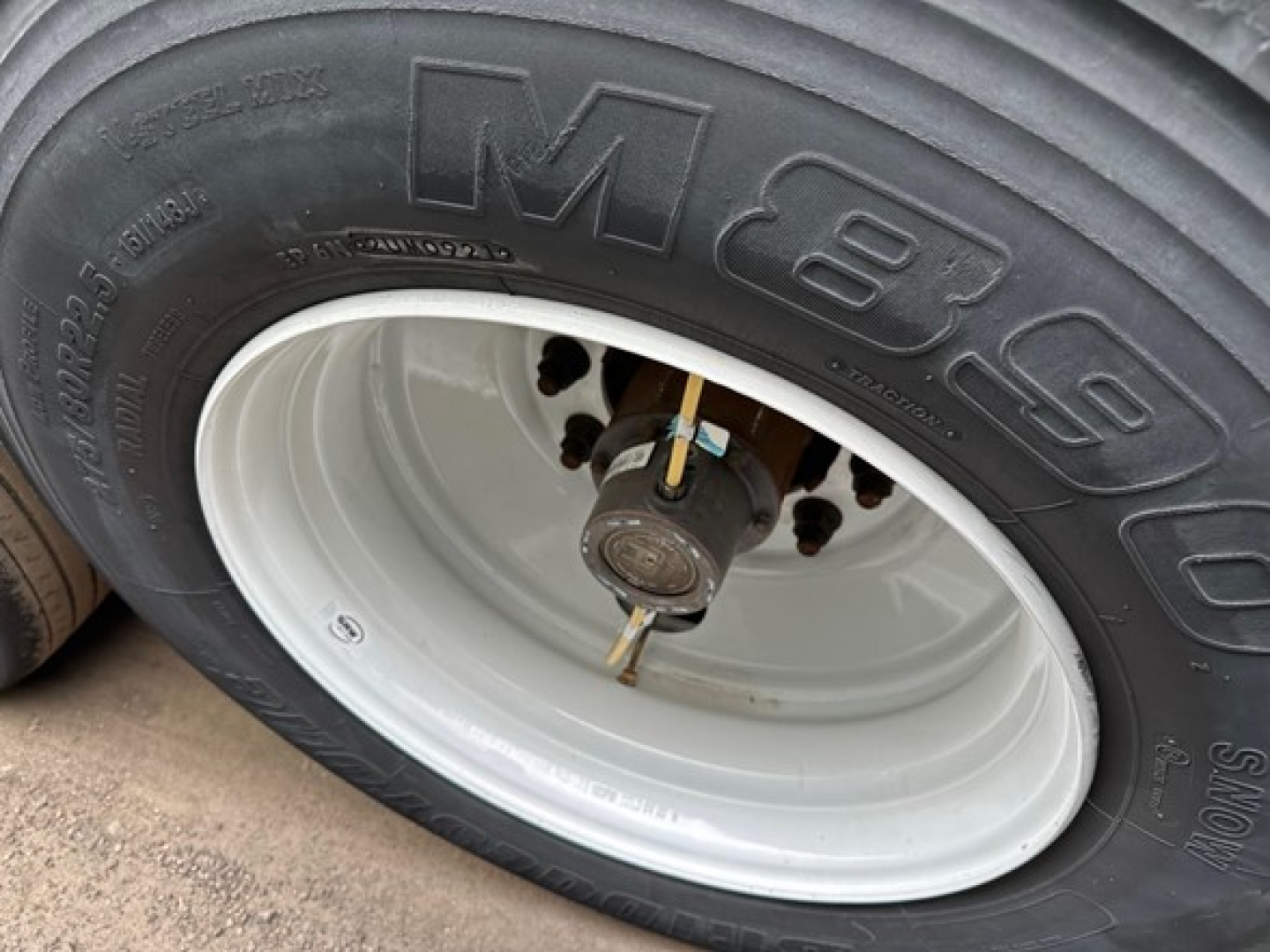 Trailer wheel close-up showing M+B 8.25R tire with white steel wheel and hub. Visible tire tread, valve stem, and wheel lug nuts.