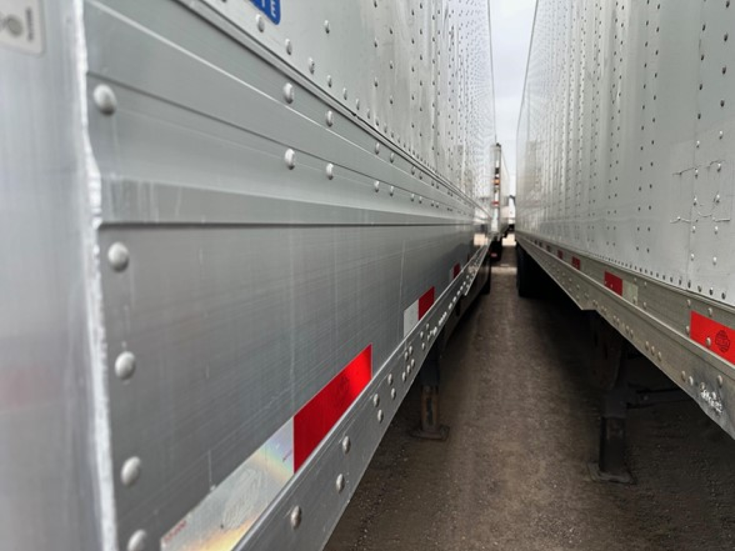 Two silver Utility trailers side-by-side with riveted metal sides and red/white reflective markings, viewed from between the trailers.