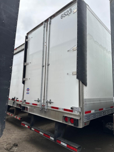 Trailer rear view showing double rear doors, silver aluminum body, black corner protectors, red and white reflective tape, and taillights.