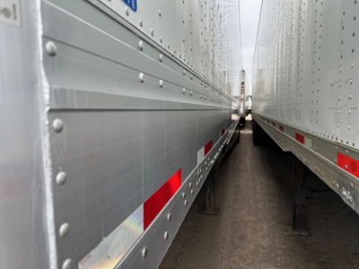 Two silver Utility trailers side-by-side with riveted metal sides and red/white reflective markings, viewed from between the trailers.