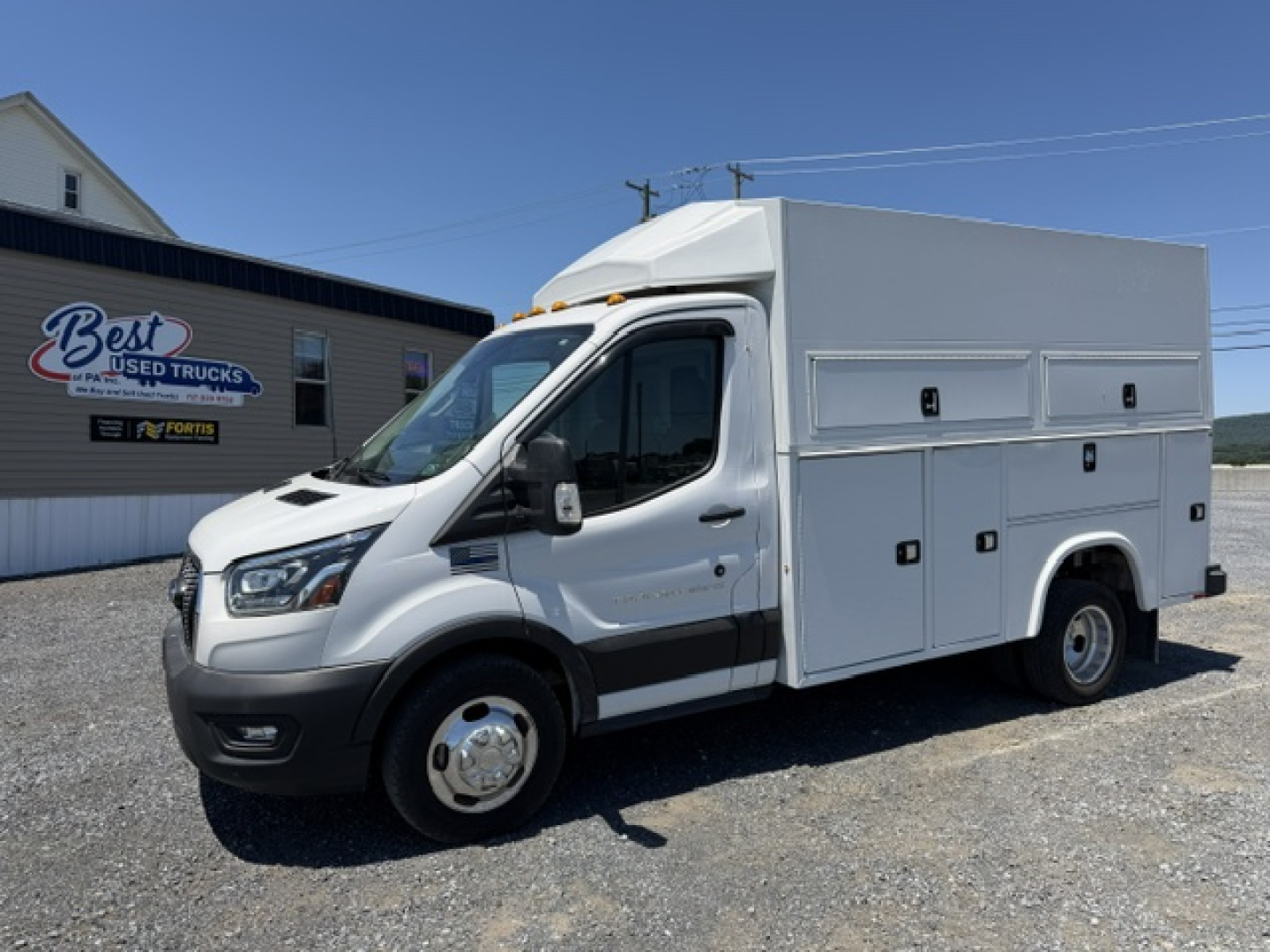 Ford Transit 350HD service vehicle in a light-gray utility body with side compartments and dual rear wheels, front grille and headlights visible.