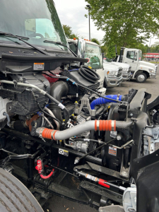Kenworth T280 engine bay view showing intake hoses, coolant reservoir, and various hydraulic lines near the front. Visible components include hoses with orange and blue couplers, radiator, and surrounding engine bay hardware.