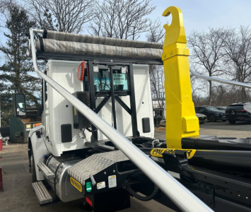 Kenworth T280 hooklift truck body showing hydraulic crane and yellow hooklift arm and side fuel tank with steps; cab rear with exhaust pipe visible.