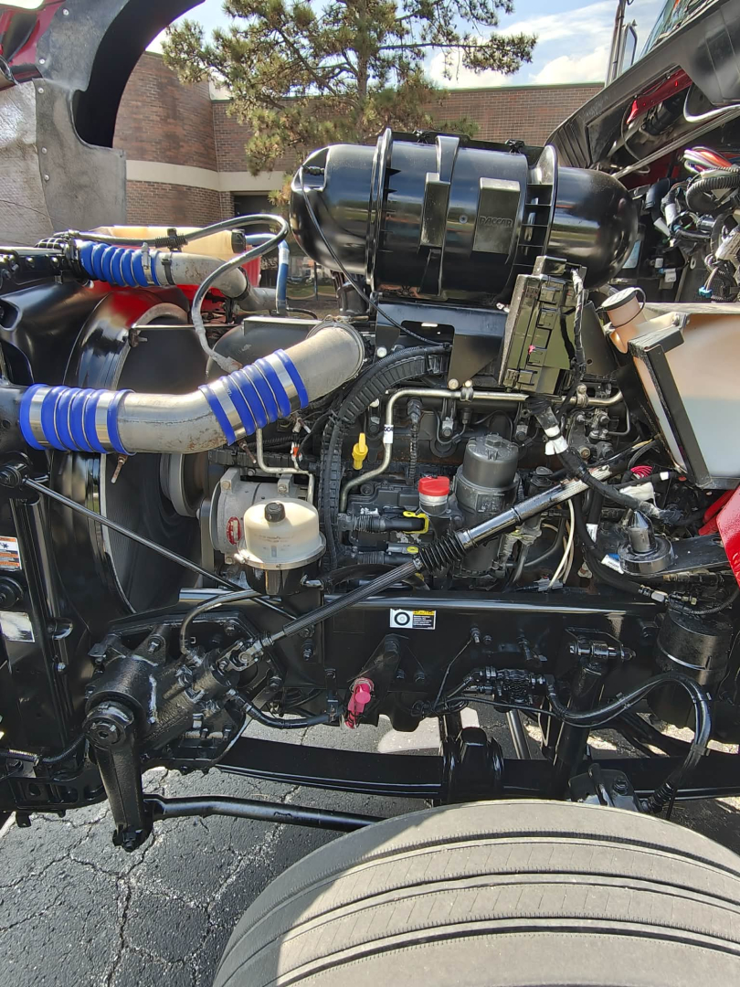 Kenworth T800 engine bay with blue, red, and silver hoses and components visible; air intake assembly, various hydraulic lines, and engine blocks visible from side view.