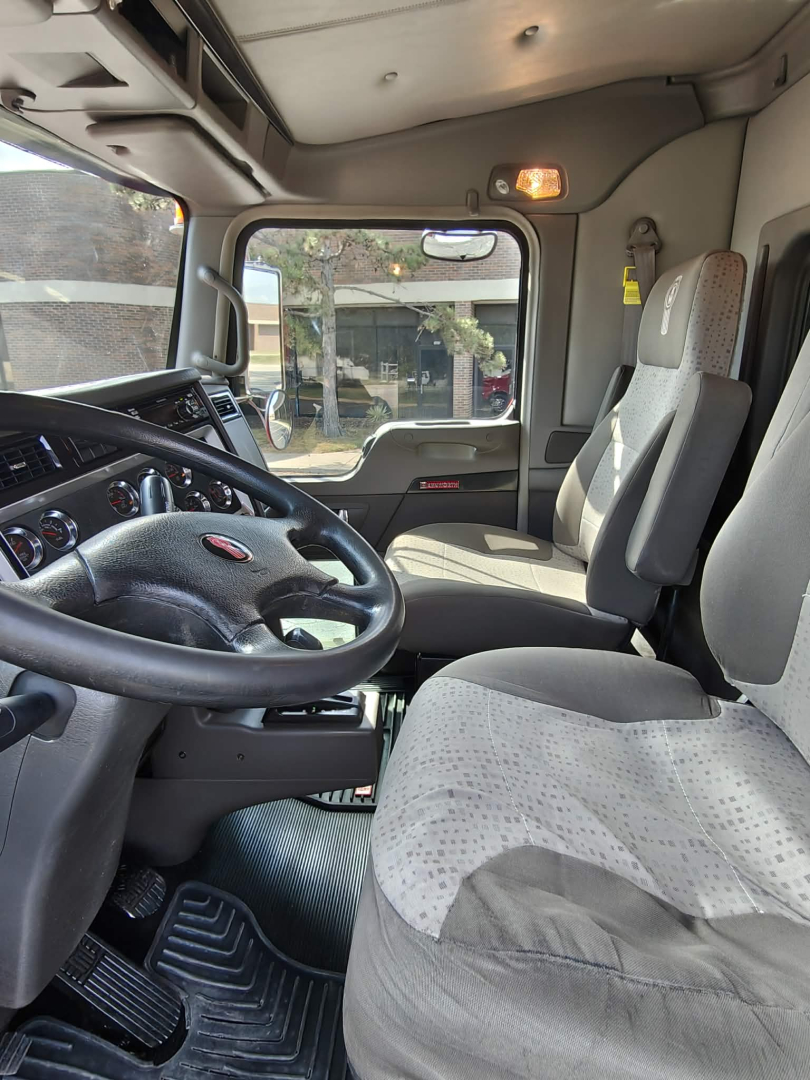 Cab interior of 2020 Kenworth T800 truck showing steering wheel, driver seat, passenger seat, dashboard, and door with window; view through windshield and side window.