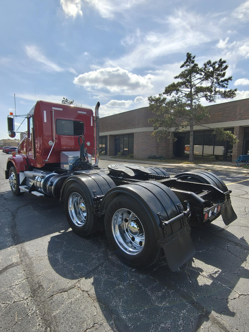 Kenworth T800 tractor in a parking lot, rear view showing dual rear axles and chrome wheels, red cab, fuel tanks, and fifth wheel in clear view.