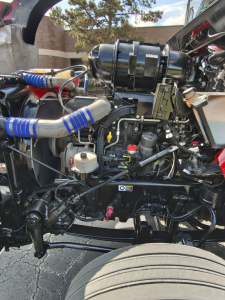 Kenworth T800 engine bay with blue, red, and silver hoses and components visible; air intake assembly, various hydraulic lines, and engine blocks visible from side view.