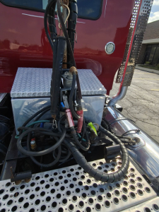 Kenworth T800 engine bay area with metal perforated step and chrome exhaust/air lines, silver toolbox, and various hoses and cables around the cab.