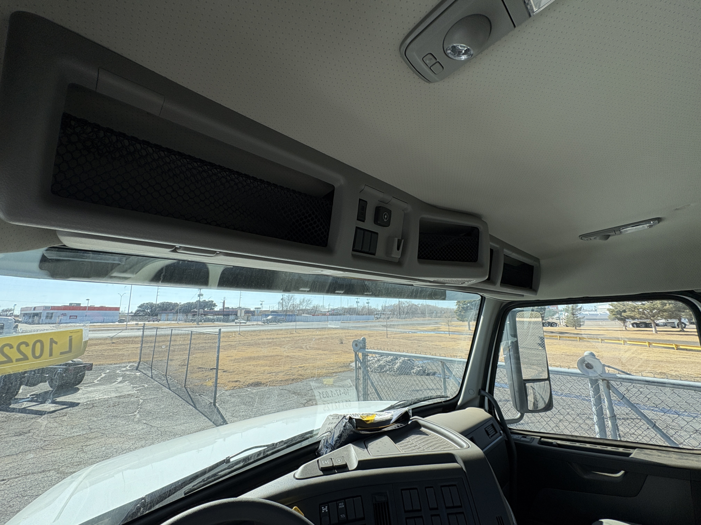 Interior cab view of a Volvo VHD truck showing overhead dash panels, storage compartments, and sun visor; windshield with exterior lot visible.