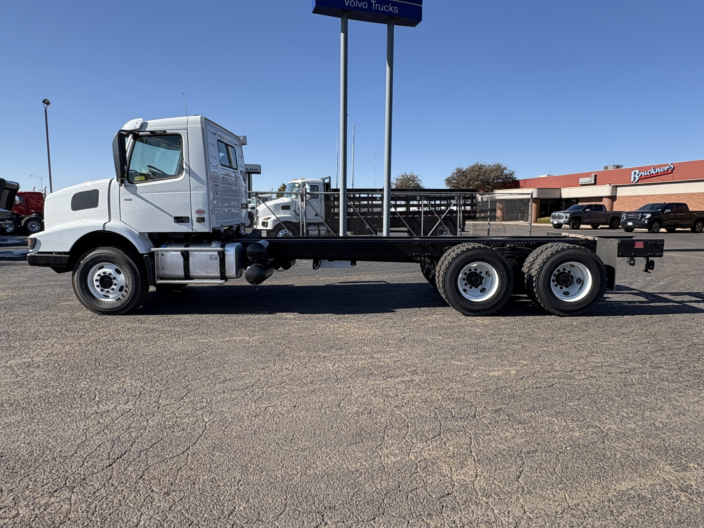 Side view of a white Volvo VHD64B300 truck chassis with long flatbed trailer and six rear wheels, in a dealership lot.
