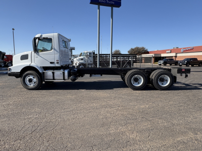 Side view of a white Volvo VHD64B300 truck chassis with long flatbed trailer and six rear wheels, in a dealership lot.