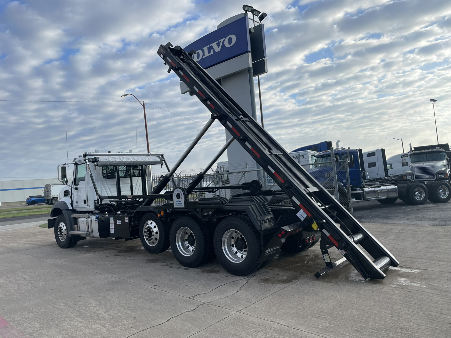 New 2026 Mack GR64B roll-off truck in a dealership lot with loading ramps raised, three-axle rear, white cab, black chassis, and Volvo sign in background.