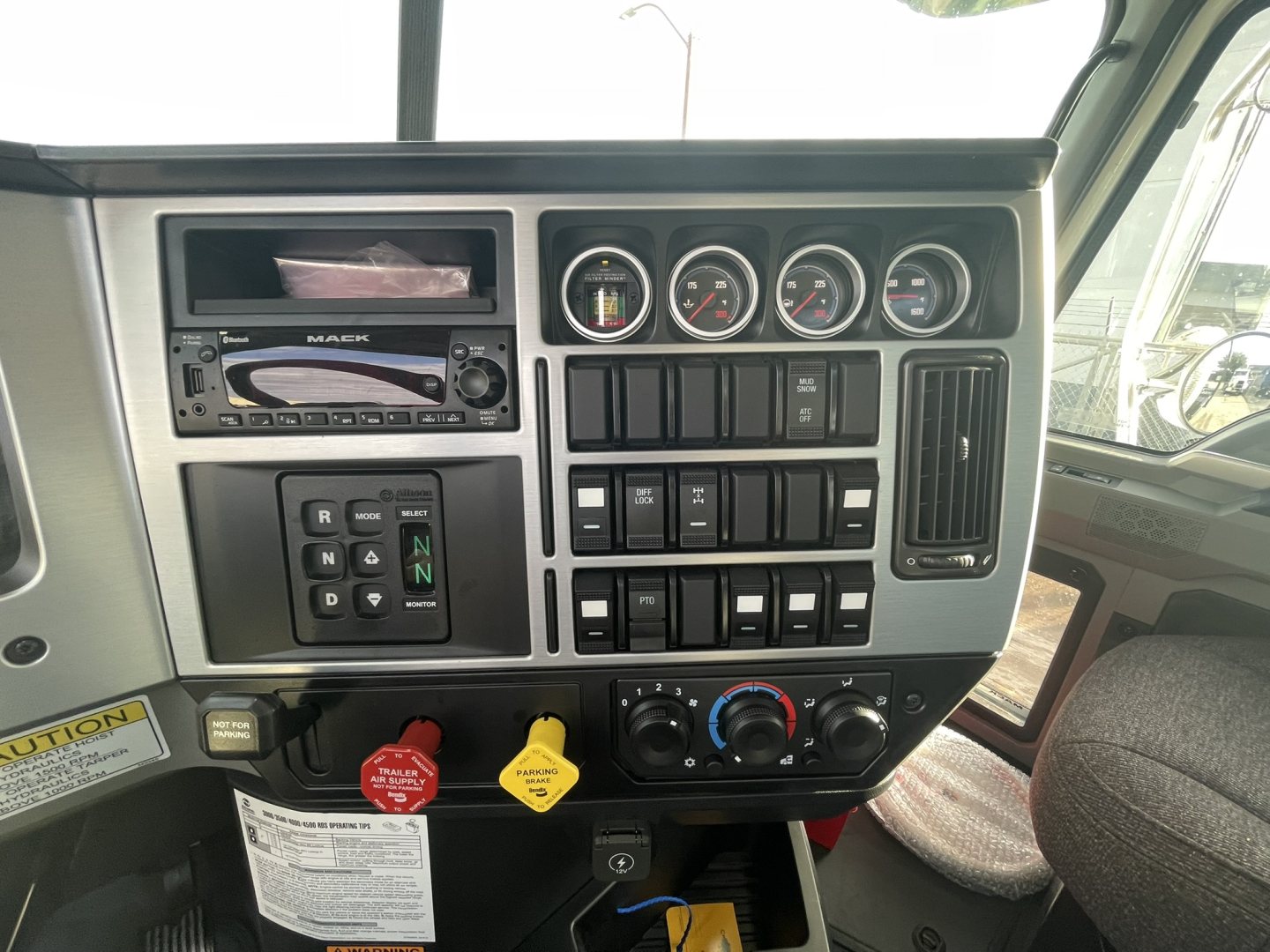 Dashboard and control panel of a Mack GR64B truck showing gauges, switches, and ignition area with red and yellow handles.