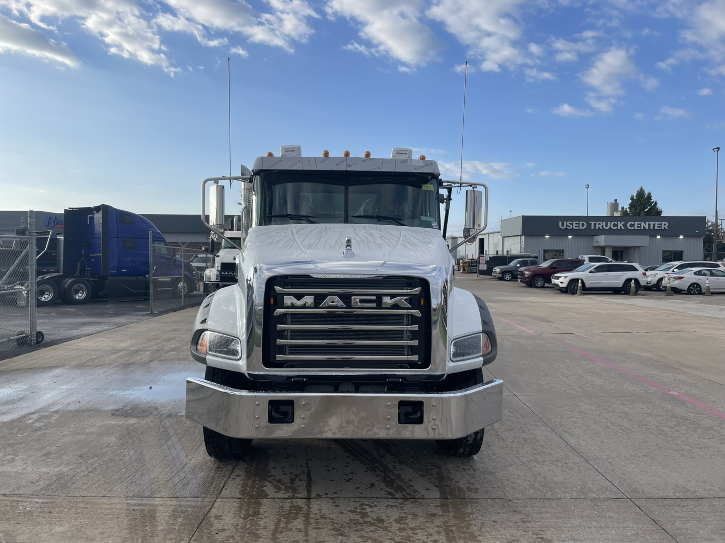Front view of a white Mack GR64B truck with large grille, bumper, and dual mirrors, on a dealership lot.