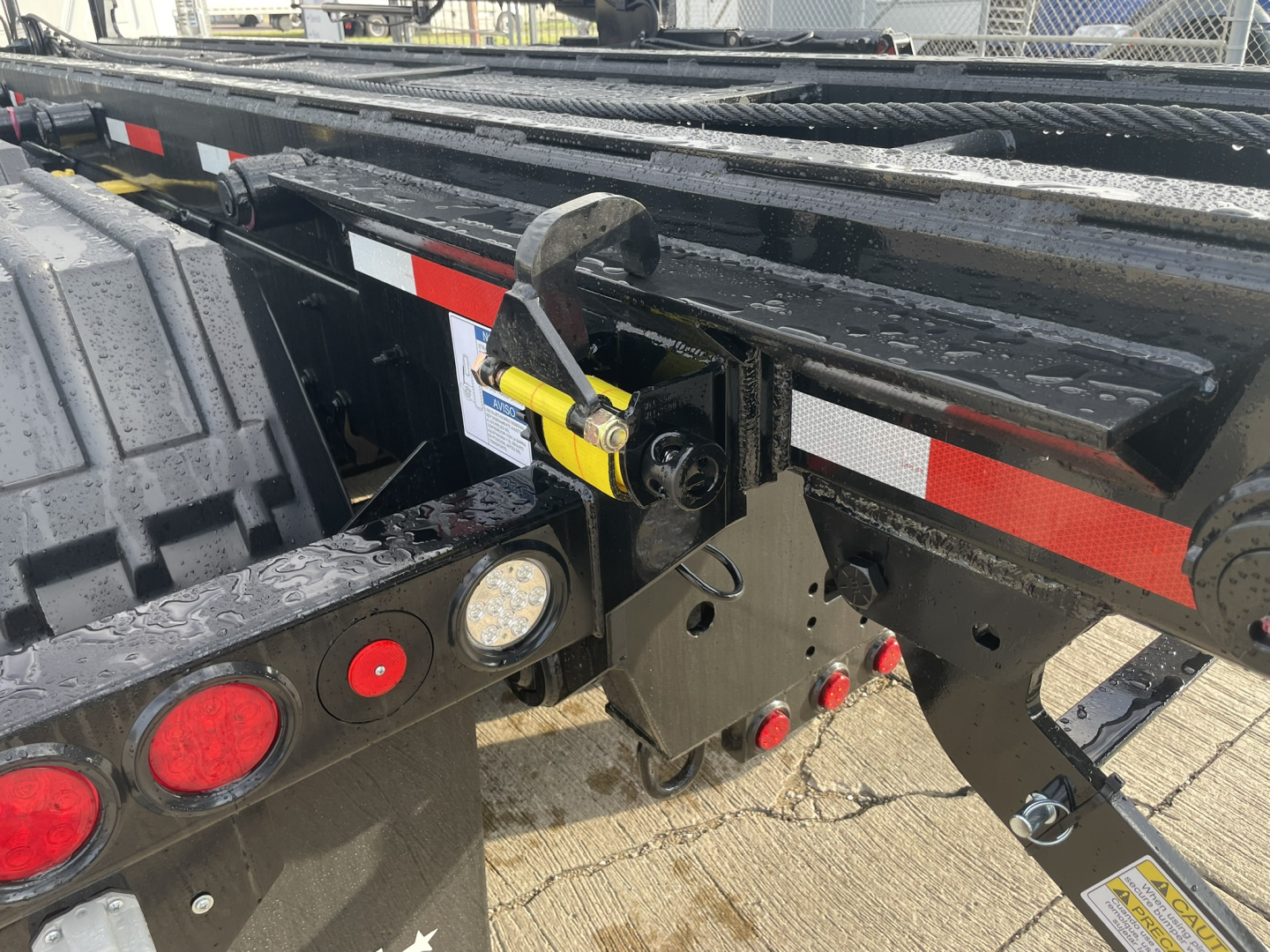 Mack GR64B roll-off truck rear chassis showing hydraulic latch assembly and safety chain hook, red/white reflective tape, and circular taillights.