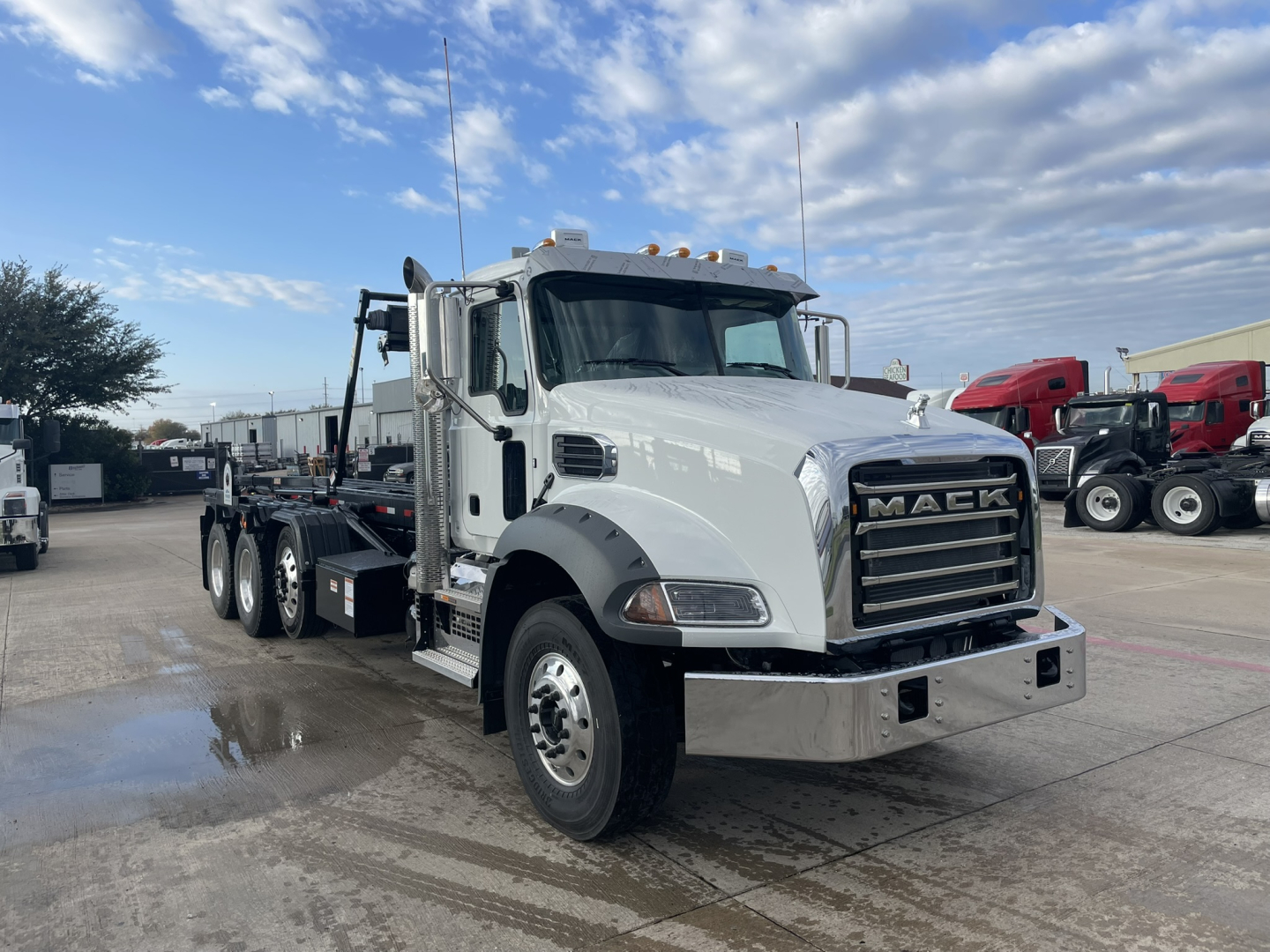 Mack GR64B roll-off truck in a dealership lot, front three-quarter view showing gray cab, heavy-duty bumper, chrome grille with MAEK letters, and a long wheelbase chassis.