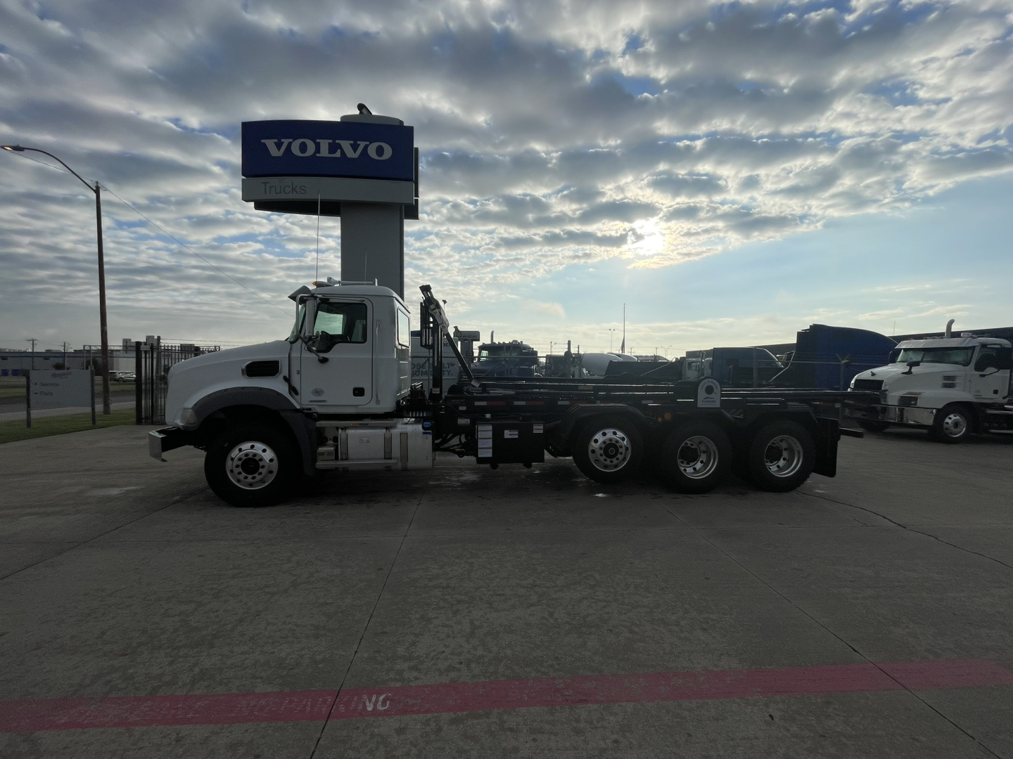 2026 Mack GR64B roll-off truck with long wheelbase, white cab, multiple axles, and flatbed cargo area in a dealership lot.