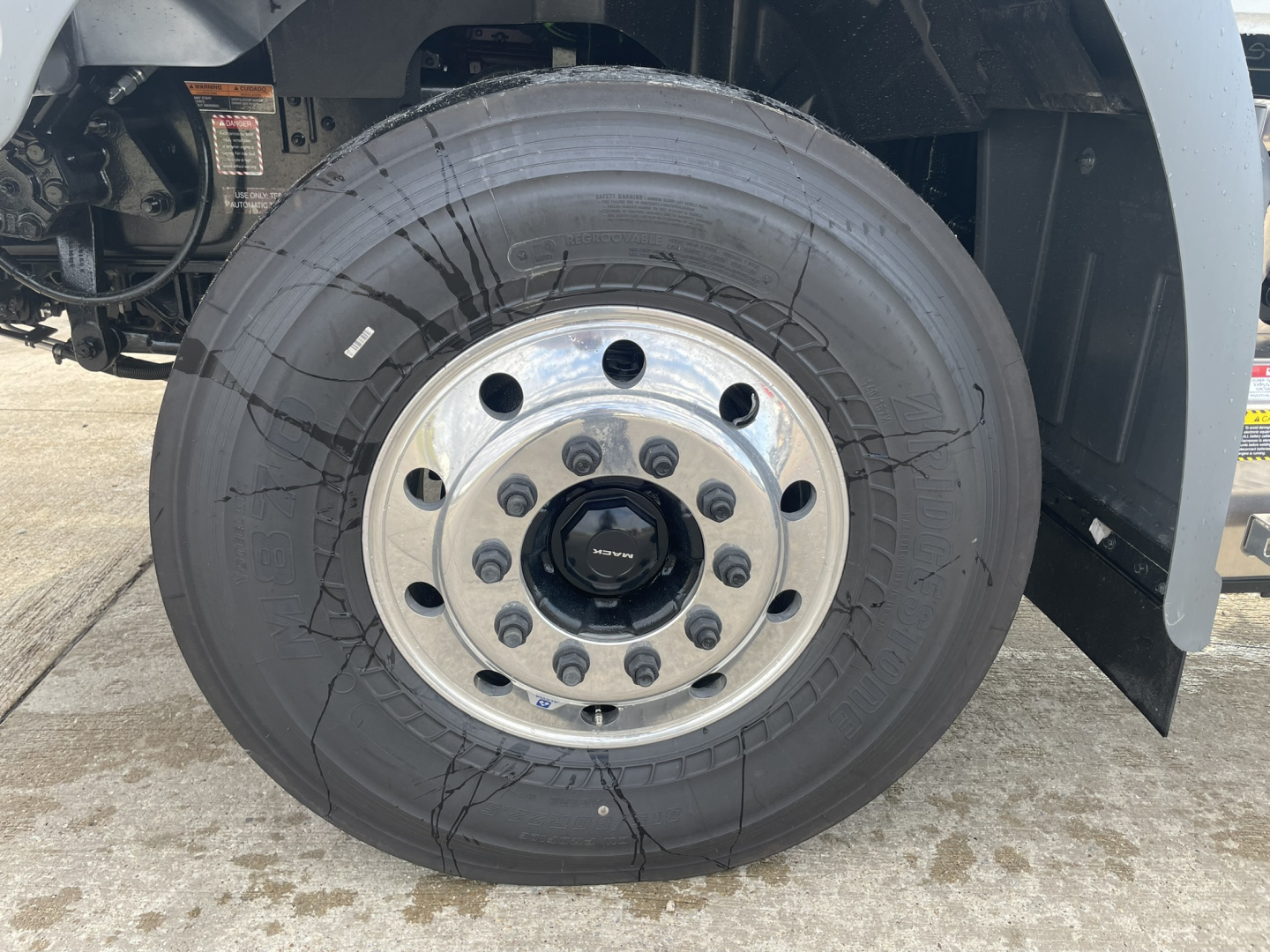 Wheel and tire on a Mack GR64B front axle showing hub with multiple lug nuts and center cap. No visible text readable on tire sidewall.