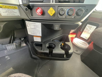 Mack GR64B cab interior showing control panel with red engine start button, yellow caution label, gauges, and cup holders.