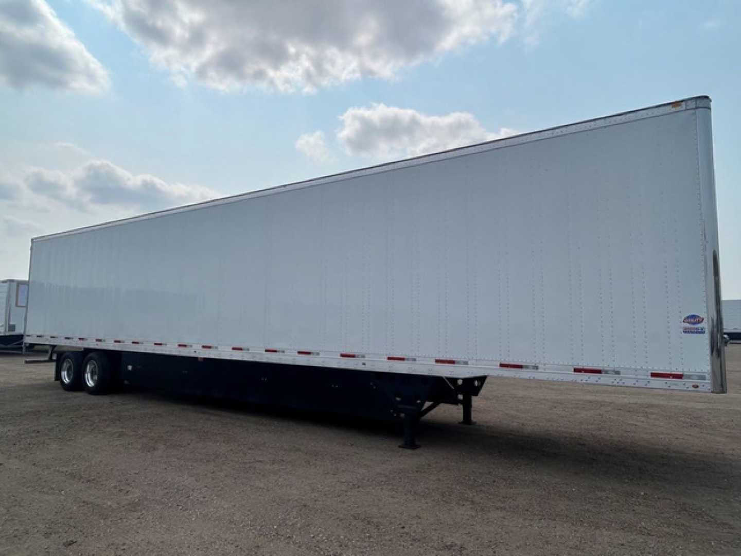 Trailer in a parking lot showing a long white box-style utility trailer with dual rear axles and a front hitch, no visible branding.