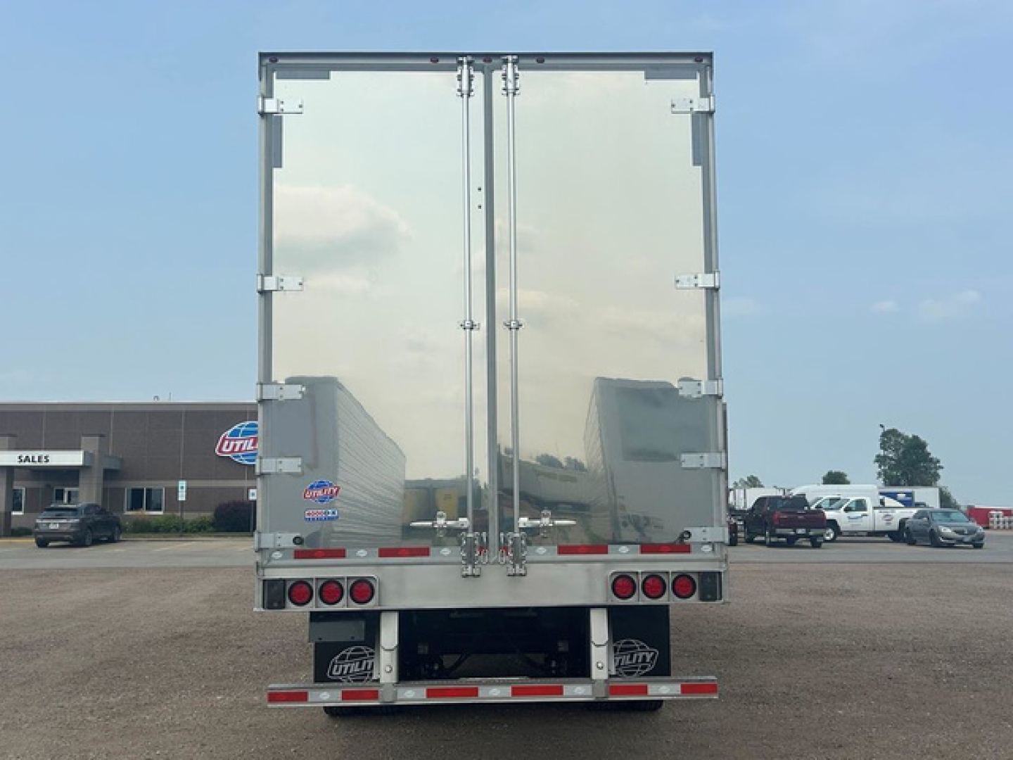 Rear view of a new Utility Dx trailer with reflective silver doors and rear bumper, in a dealership lot.