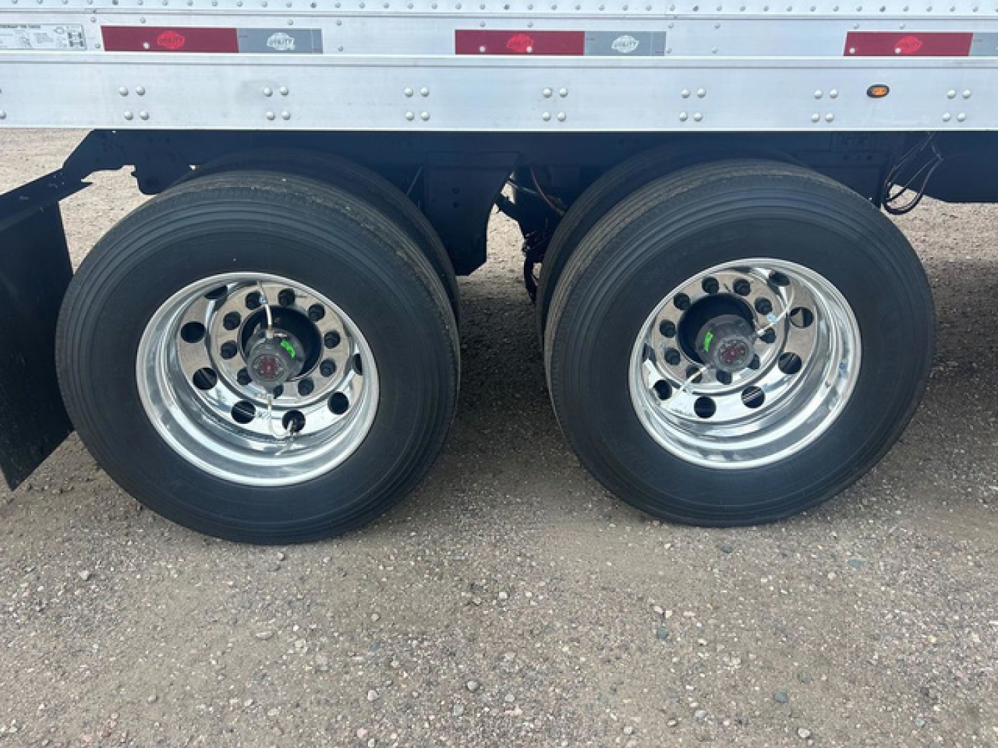 Trailer axle pair showing dual rear wheels on a Utility Dx trailer in new condition. Rust-free chrome rims with black tires, undercarriage and reflective tape visible.