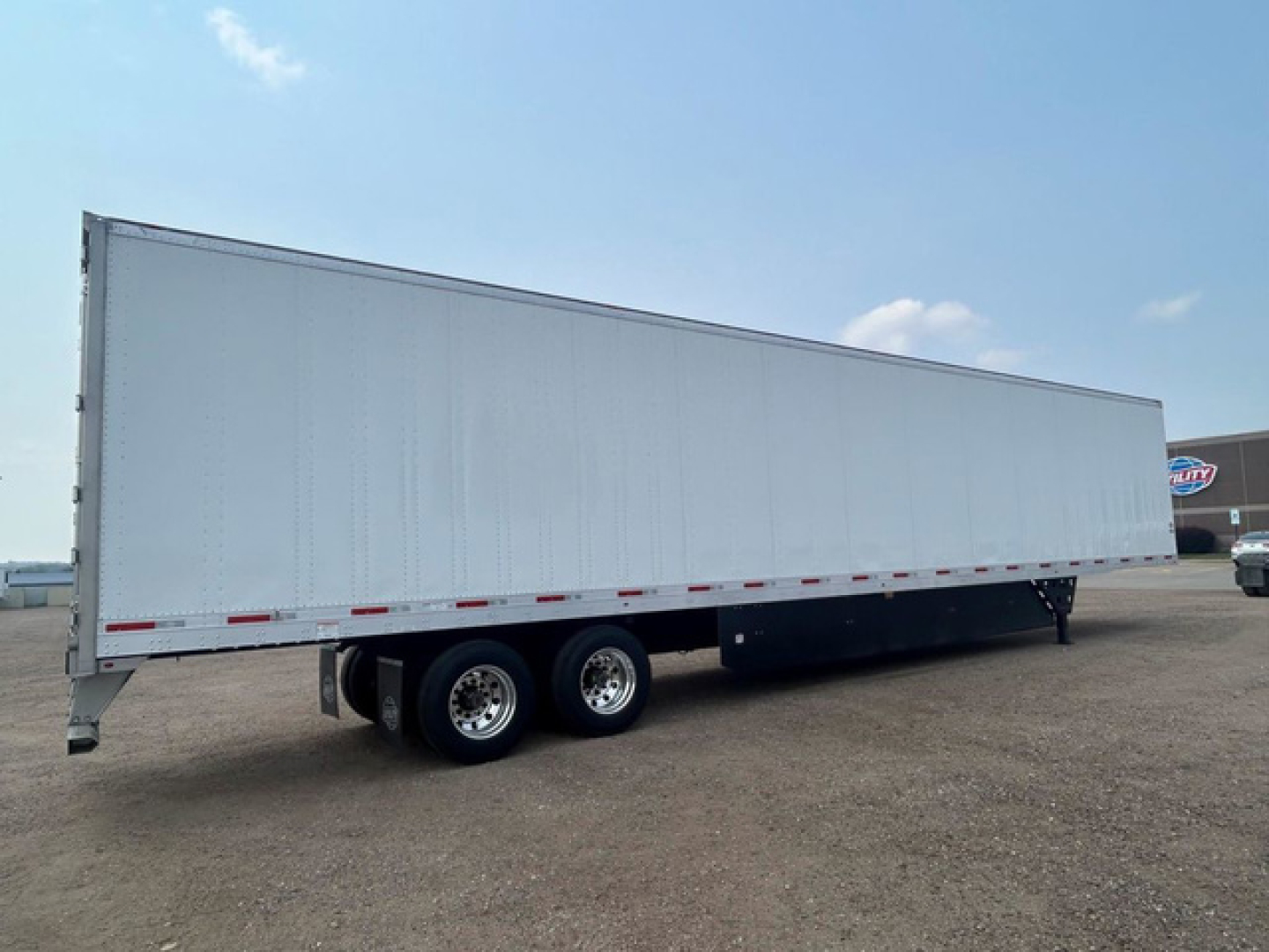 Utility Dx trailer in a dirt lot with tandem axle and white exterior, side view showing long cargo area and red/white reflective tape along the lower edge.