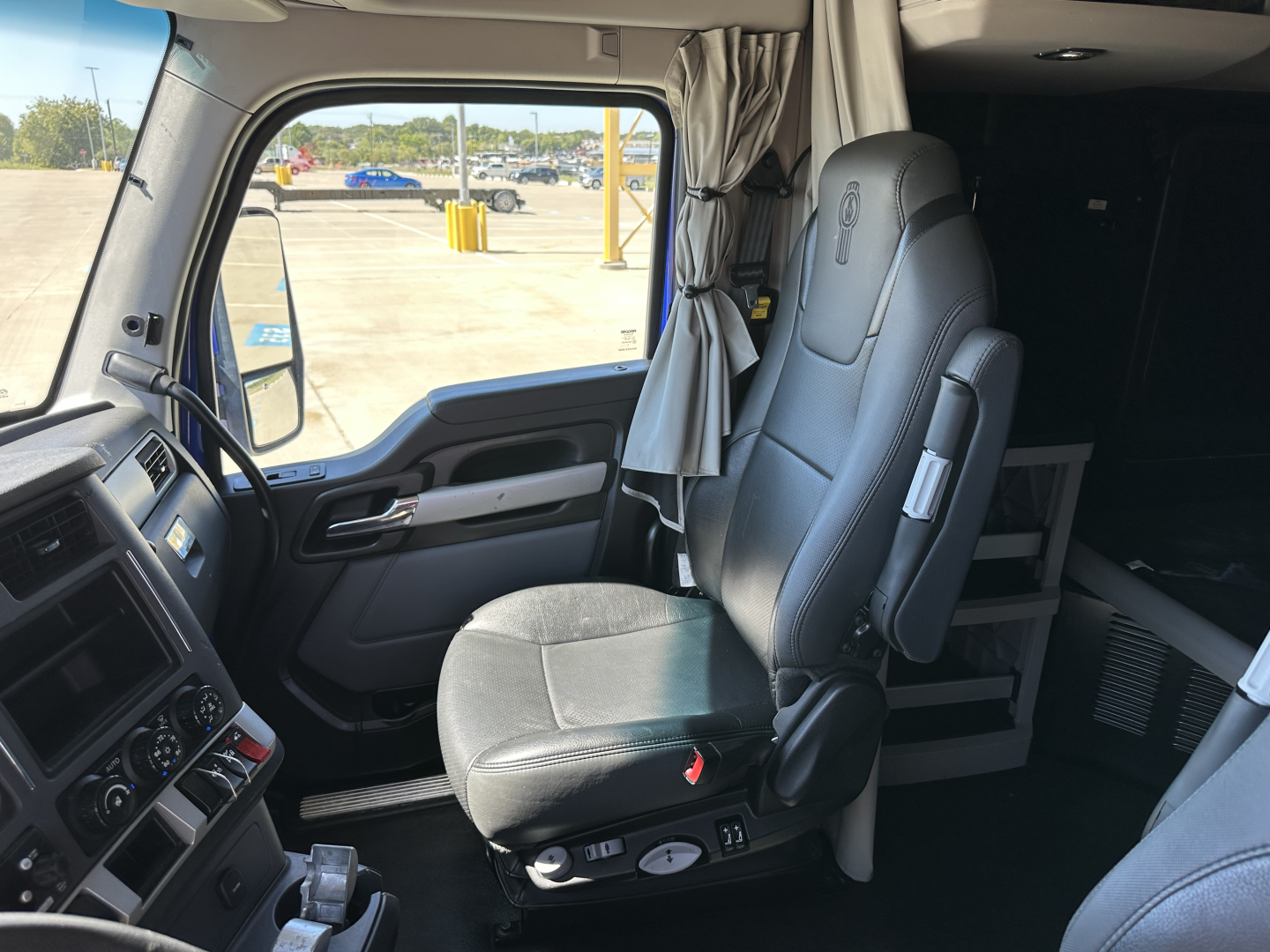 Truck cab interior of a 2023 Kenworth T680 showing driver seat and steering wheel with dashboard and window, light gray seating and curtains.