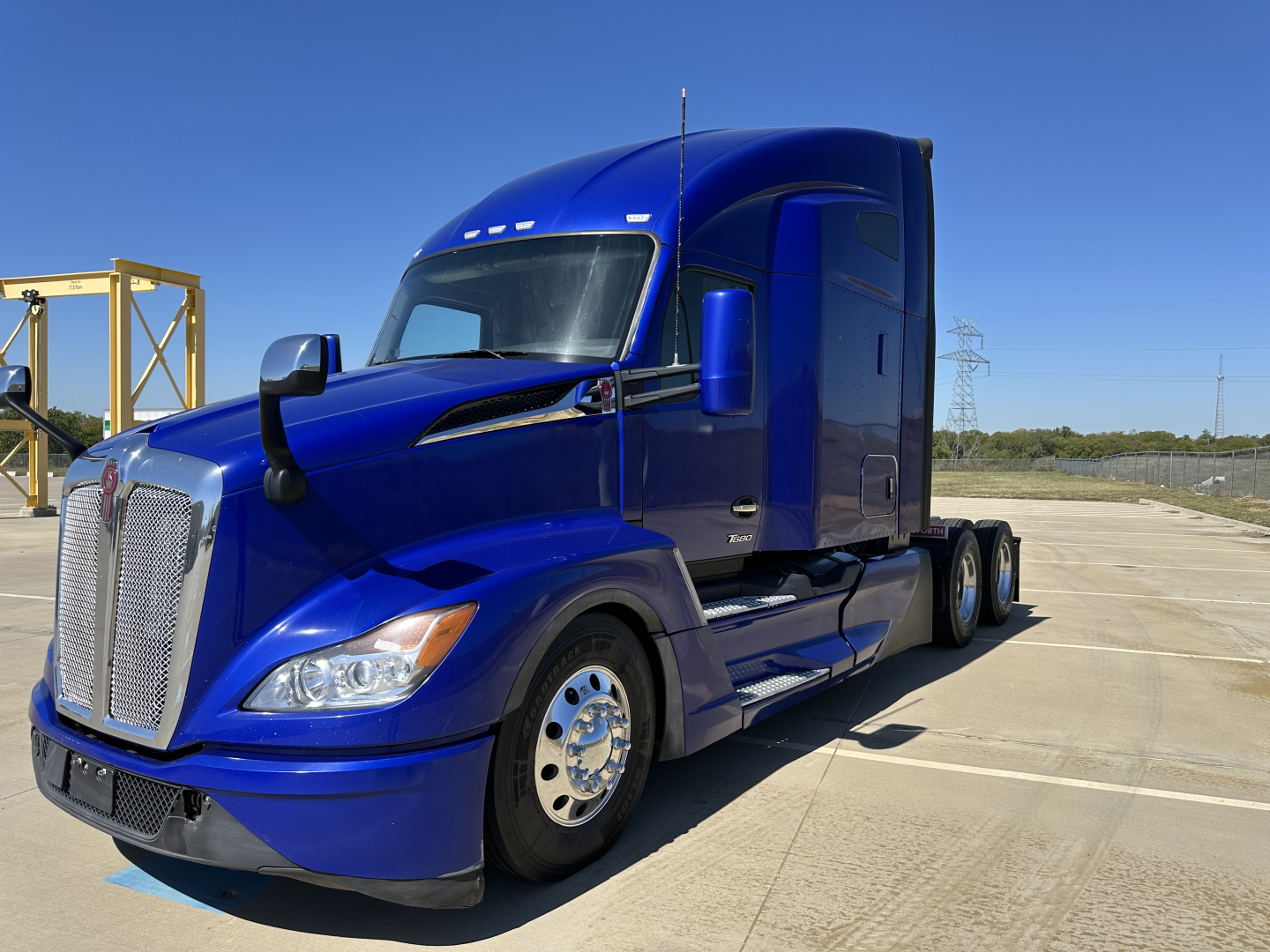 Blue 2023 Kenworth T680 truck in a parking lot, facing left, with triple-axle rear, chrome wheels, and large grille.