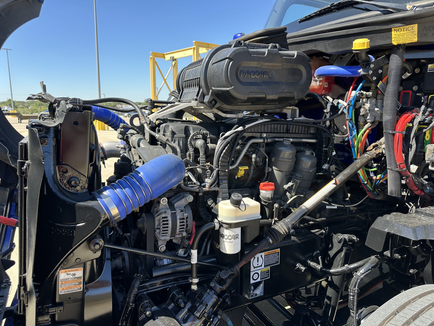 Kenworth T680 engine bay showing Cummins/Isuzu style components, hoses, reservoirs, and electrical wiring under hood.
