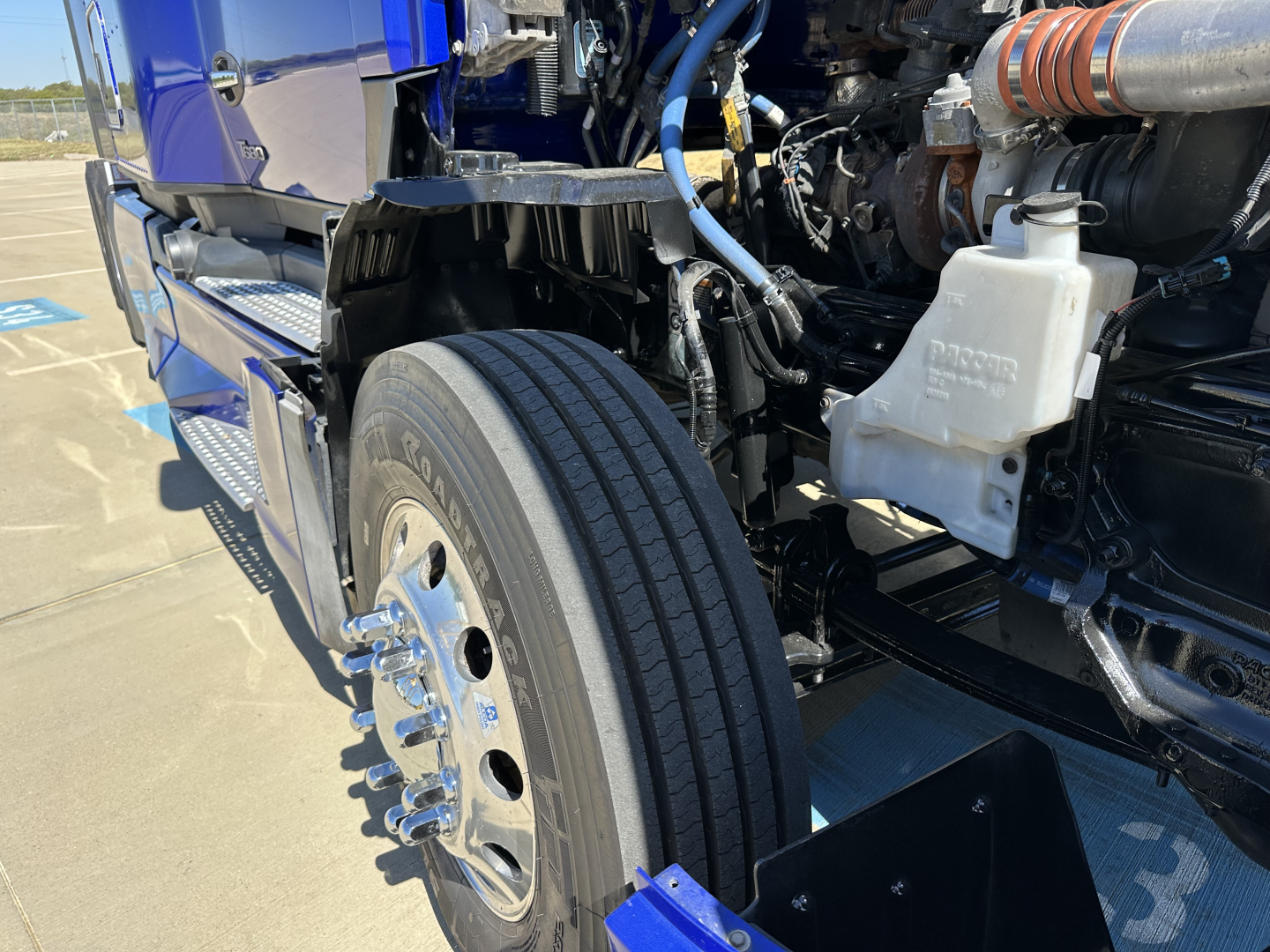 Kenworth T680 engine bay with front left wheel visible, showing coolant reservoir, air intake duct, and various hoses and metal components.