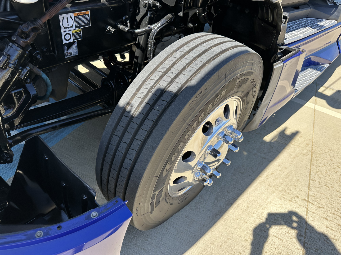 Truck front wheel and suspension close-up showing steering hub and tire on Kenworth T680 chassis with metallic wheel, bolts, and black undercarriage components.