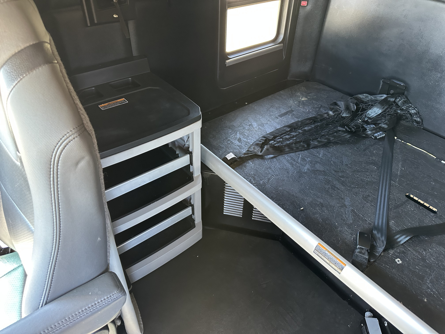 Kenworth T680 interior view showing sleeper area with seat, storage drawers, and seat belt harness.