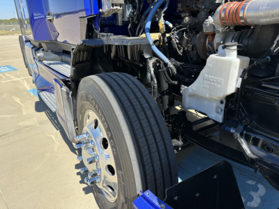 Kenworth T680 engine bay with front left wheel visible, showing coolant reservoir, air intake duct, and various hoses and metal components.