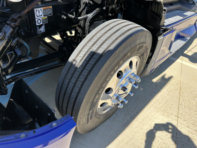 Truck front wheel and suspension close-up showing steering hub and tire on Kenworth T680 chassis with metallic wheel, bolts, and black undercarriage components.