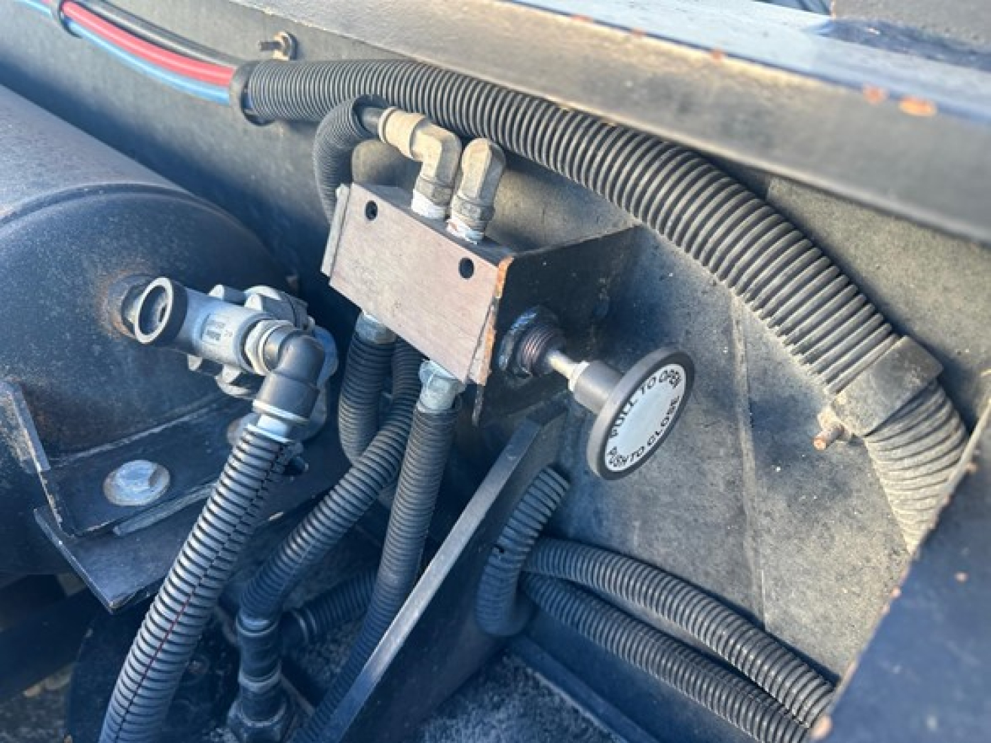 Trailer wiring and hydraulic component in engine bay of Fontaine lowboy trailer showing metal block with hydraulic lines and circular cap, braided wiring loom, and conduits.