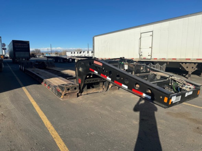 Trailer frame and lowboy with metal beams and hydraulic components, side rails and reflective striping visible in a parking lot.