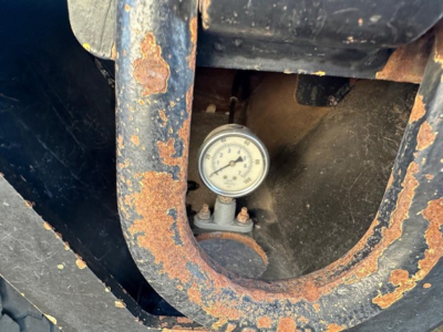 Trailer component close-up showing rusted metal frame and a dial gauge inside a circular cutout.