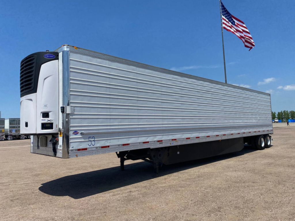 Utility refrigerated trailer with a white Thermo King unit on the front, silver corrugated side panels, tandem rear axles, and a flag in the background.