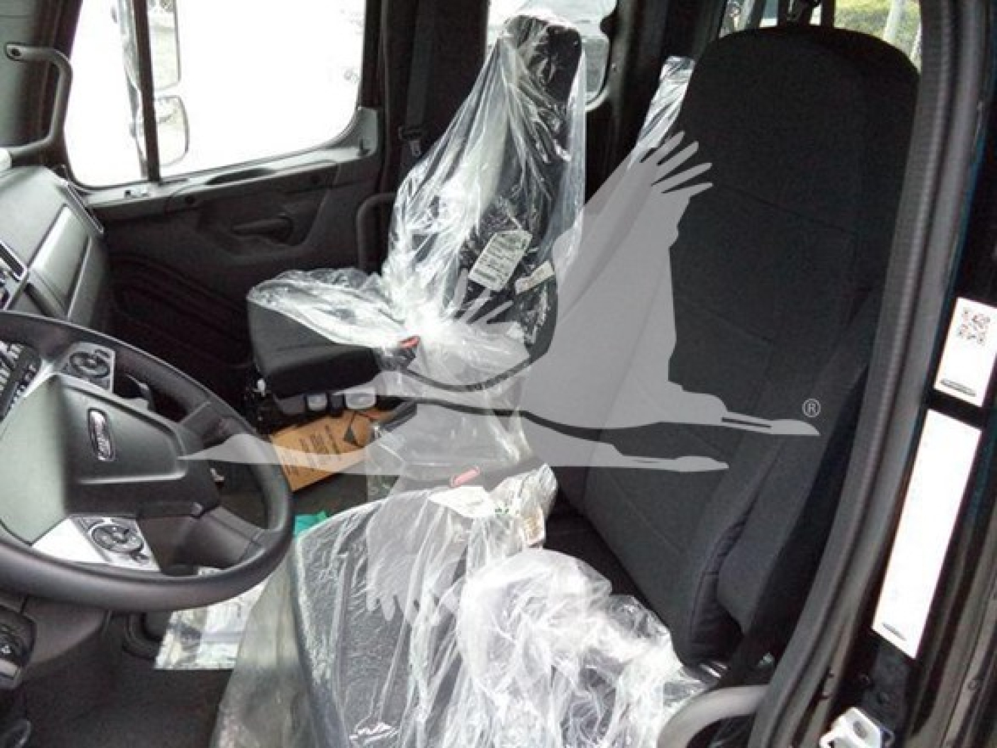 Truck interior of a 2025 Freightliner M2 106 showing driver seat and front passenger seat wrapped in plastic, steering wheel, dashboard, and door frame.