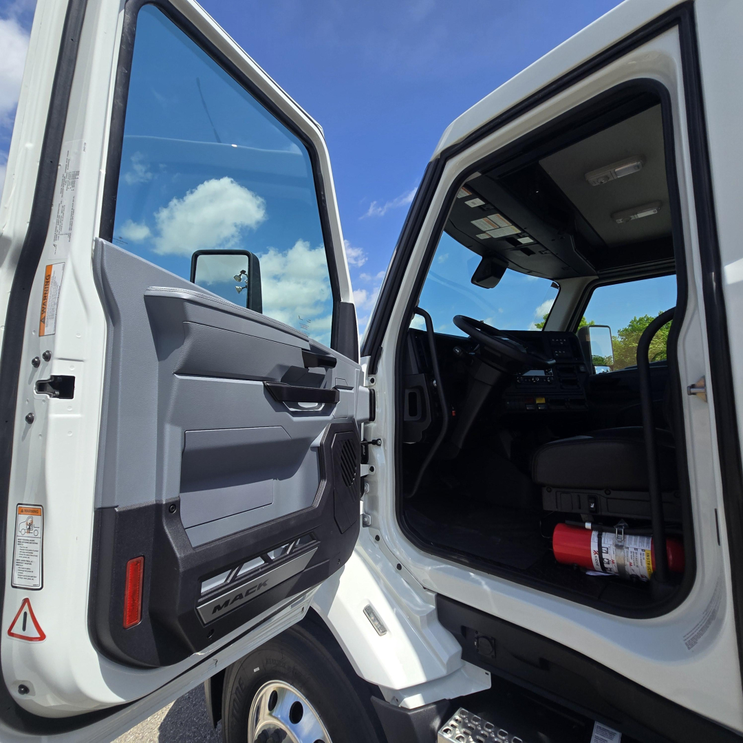 Truck cab interior view through open door of a white Mack AN64T daycab showing driver seat, dashboard, and steering wheel with a fire extinguisher on the floor.