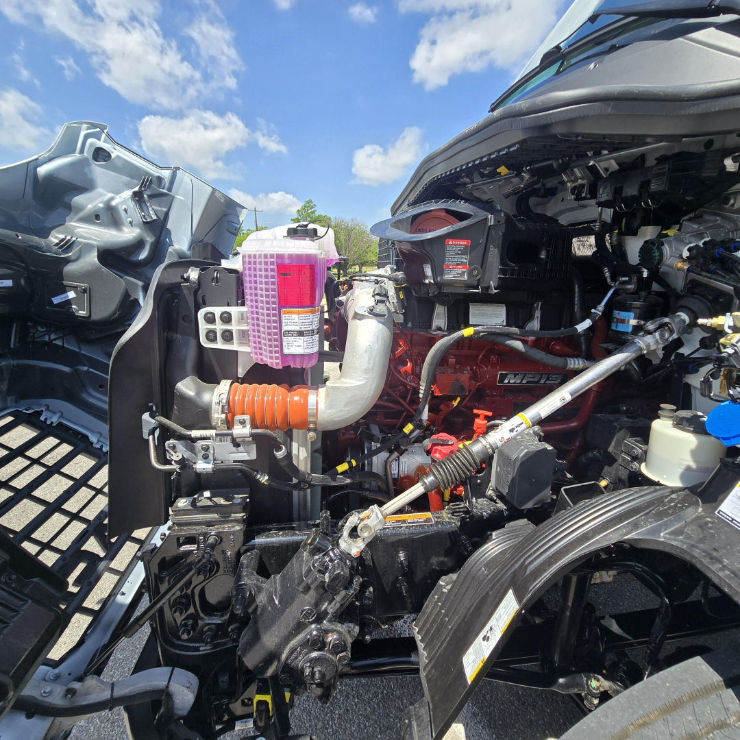 Truck engine bay with pink coolant reservoir, orange air intake hose, various hoses and metal components; visible Mack AN64T engine block in red; fender and dashboard components at right.