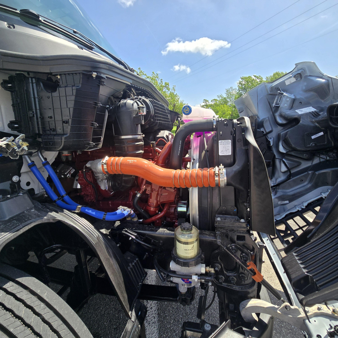 Truck engine bay showing orange coolant hose, black radiator, blue braided hose, and various engine components in a Mack AN64T cab-over-engine design.